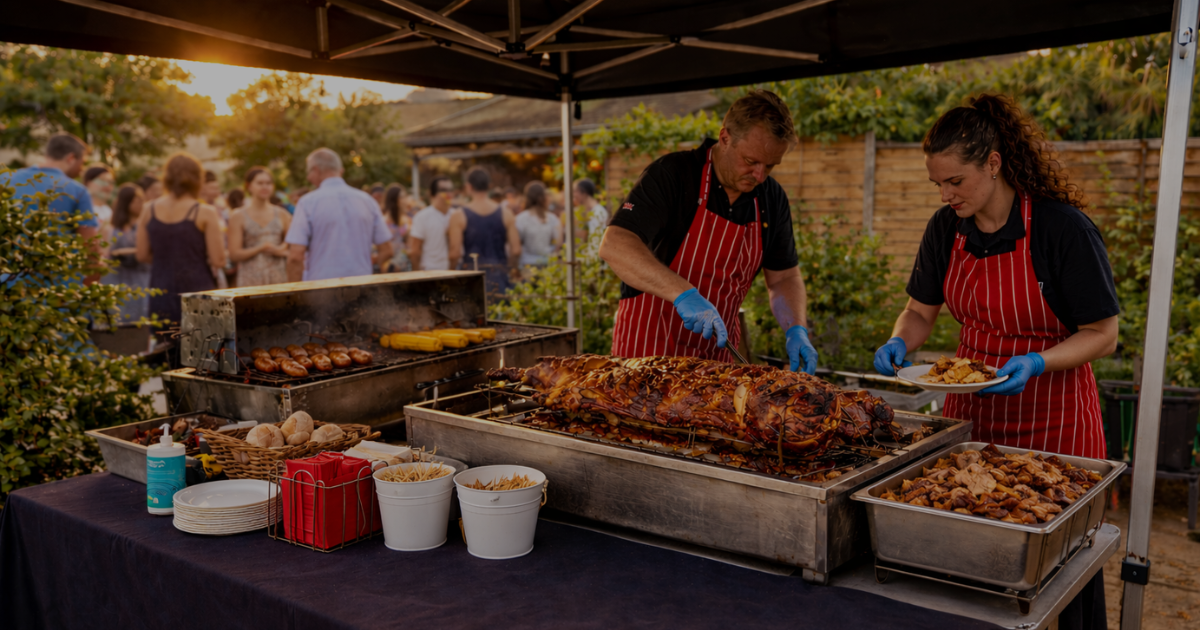 Outdoor hog roast catering setup at a summer garden event, with chefs serving freshly cooked food beside a whole roasted hog and BBQ grill while guests gather in the background at sunset.